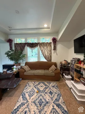 Bright living room featuring a coffered ceiling with recessed lighting, triple-window arrangement with custom drapery and floral accents, dark wood-finish flooring, patterned area rug, wall-mounted flat-screen television, and built-in shelving with displa