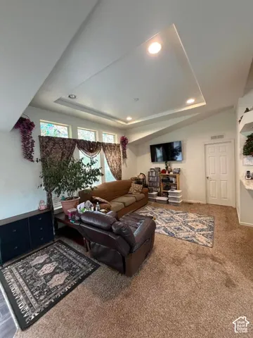 Spacious living area featuring coffered ceiling with recessed lighting, large windows with natural light, wall-mounted television, neutral carpeting, and decorative area rugs