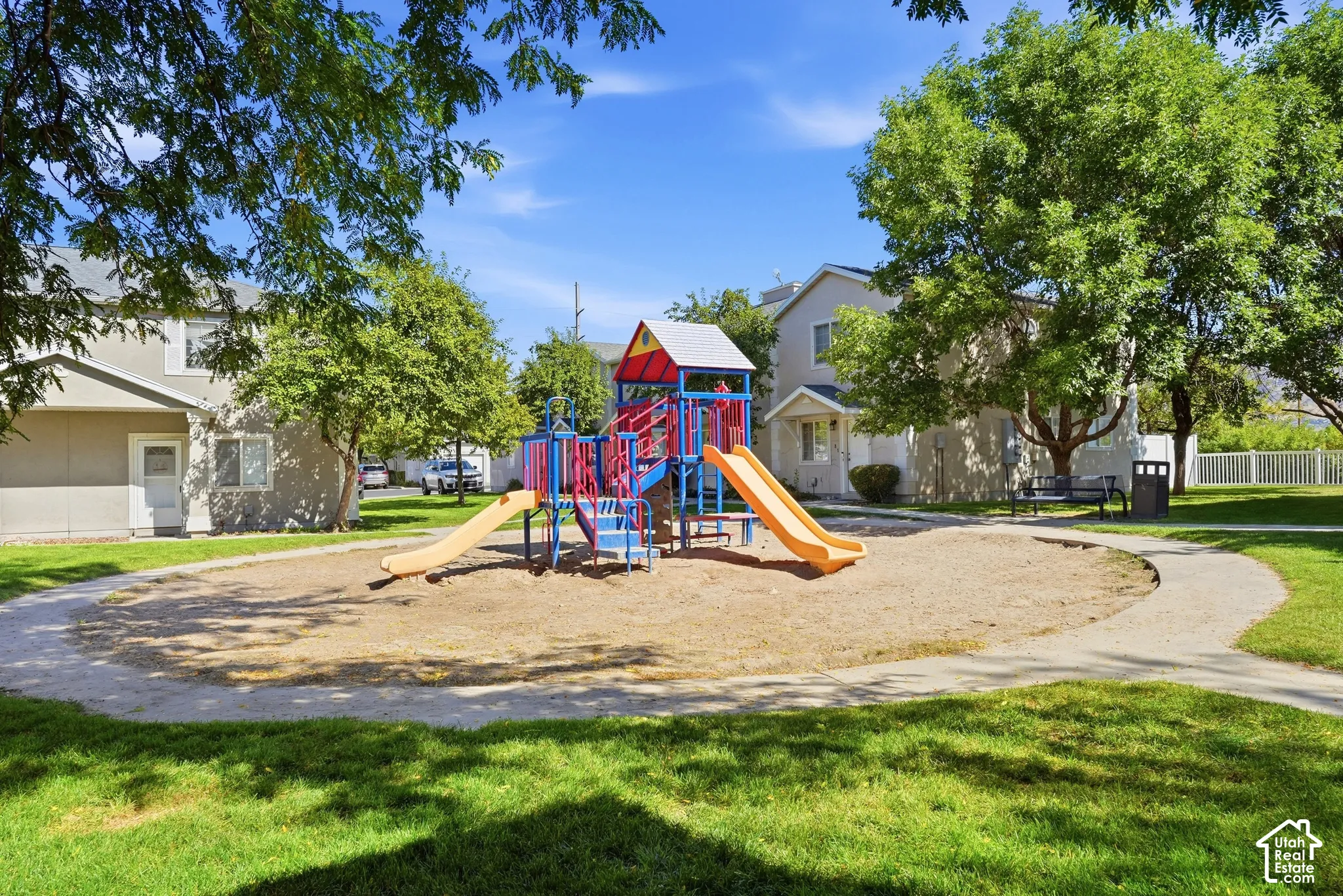 Community playground featuring slides and climbing structures, surrounded by a sand play area, manicured lawns, and mature trees