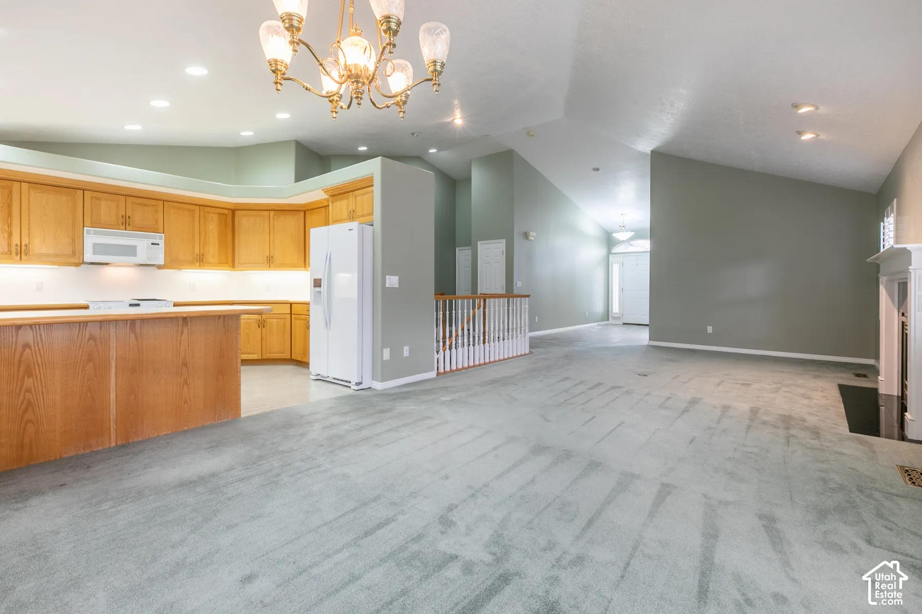 Open-concept interior featuring vaulted ceilings, natural wood cabinetry, a tiered kitchen peninsula, brass-finish chandelier, and a fireplace with a white mantel