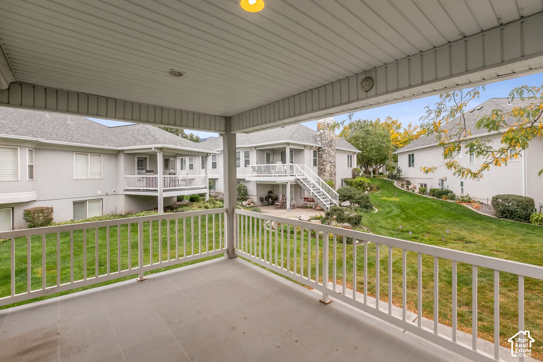 Covered balcony featuring tile flooring and white metal railing overlooking a common green space with mature landscaping