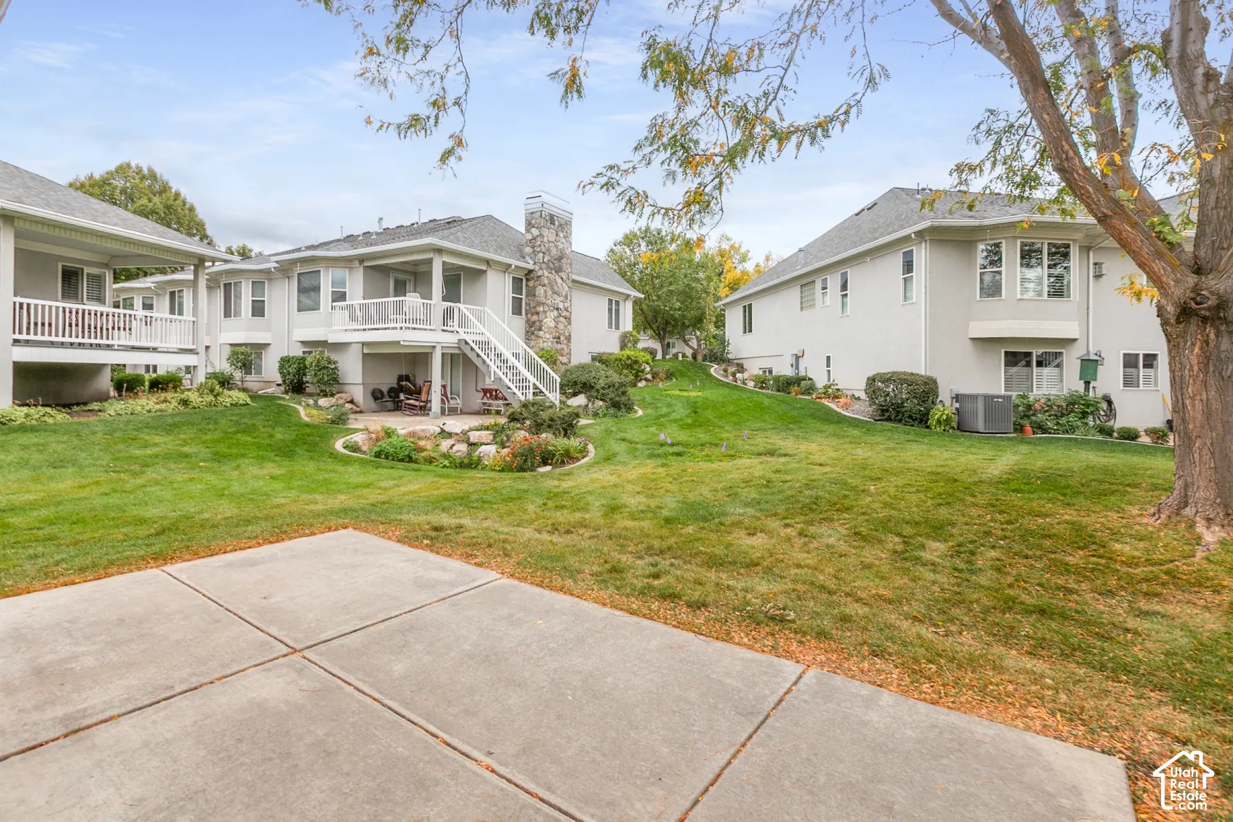 Concrete patio, elevated wood deck with white railings, stone exterior chimney, and expansive lawn with mature landscaping