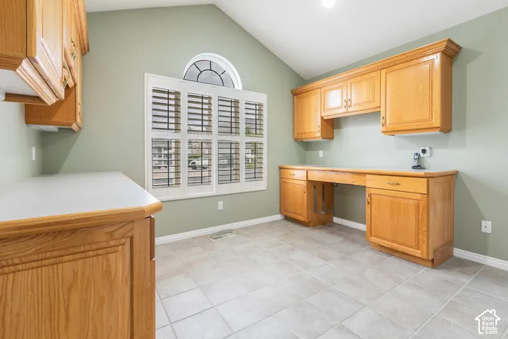 Vaulted ceiling room featuring custom wood cabinetry, extensive integrated desk space, tiled flooring, and a large window topped with a semi-circular transom