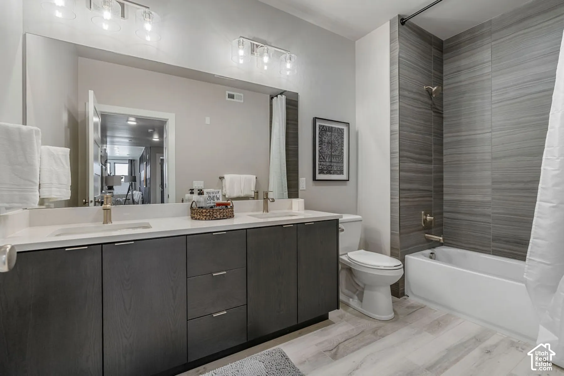 Dual vanity with light-toned countertop, wood-finish cabinetry, a large wall mirror, a bathtub with a tiled surround, and wood-finish flooring