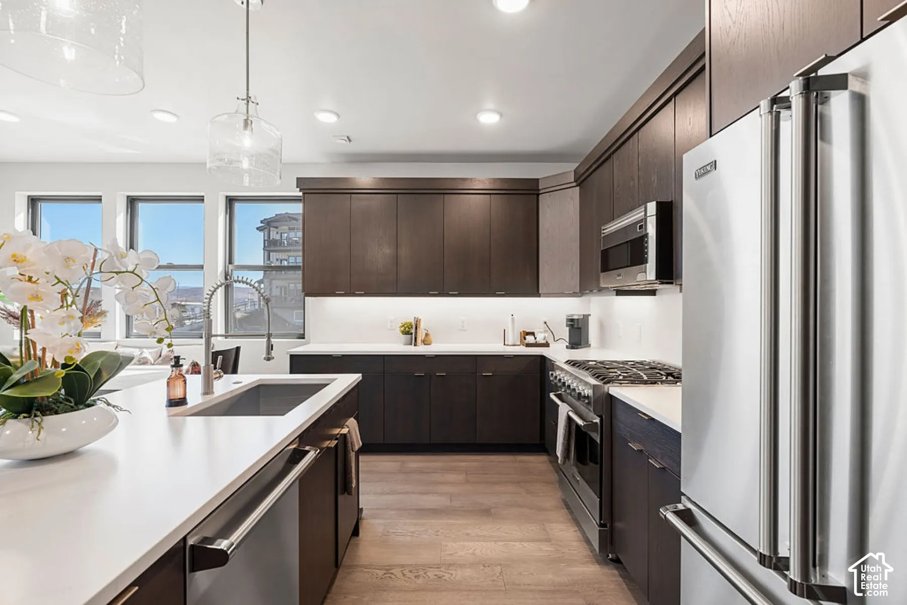 Kitchen featuring dark wood-finish cabinetry, light countertops, integrated stainless steel appliances, a stainless steel sink with a gooseneck faucet, and wood-finish flooring