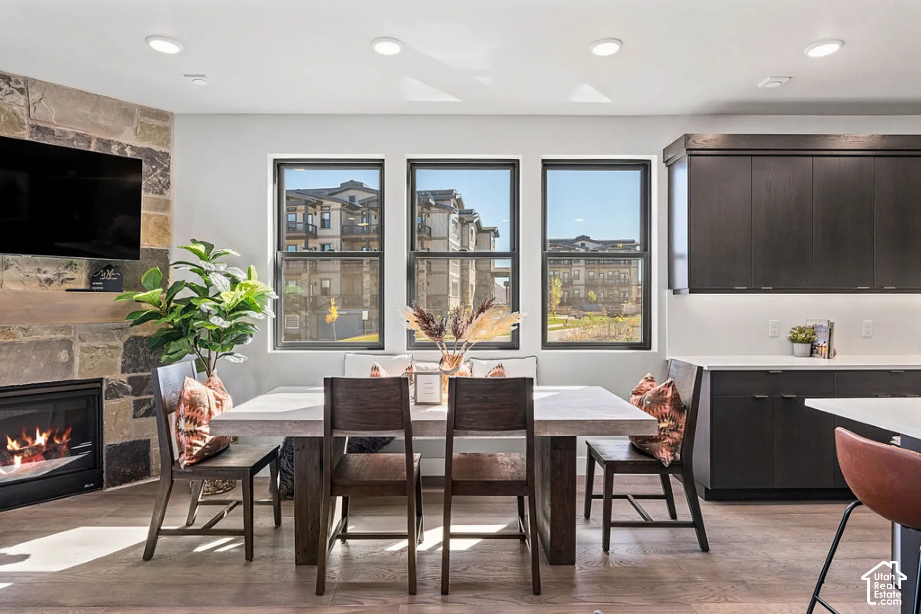 Stone-clad fireplace with a hearth, multiple black-framed windows, recessed lighting, wood-finish flooring, and dark wood-tone cabinetry