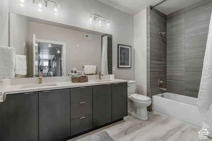 Dual vanity with light-toned countertop, wood-finish cabinetry, a large wall mirror, a bathtub with a tiled surround, and wood-finish flooring