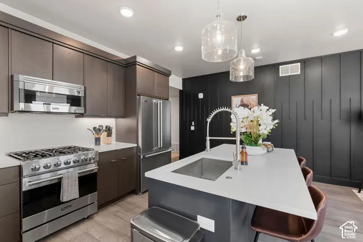 Kitchen featuring dark wood-finish cabinetry, stainless steel appliances, a center island with an integrated sink, and pendant lighting