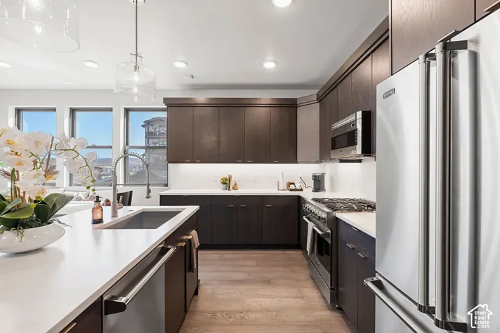 Kitchen featuring dark wood-finish cabinetry, light countertops, integrated stainless steel appliances, a stainless steel sink with a gooseneck faucet, and wood-finish flooring