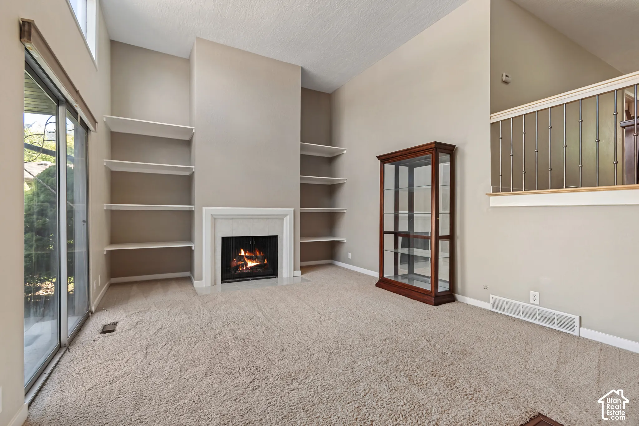 Spacious living area featuring built-in shelving on either side of a fireplace with surround, neutral carpet flooring, glass sliding doors providing natural light, and a wood-frame display cabinet with glass doors