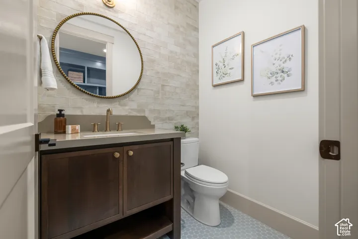 Vanity with a light-toned countertop, a gold-toned faucet, a round wall-mounted mirror, and a tile accent wall