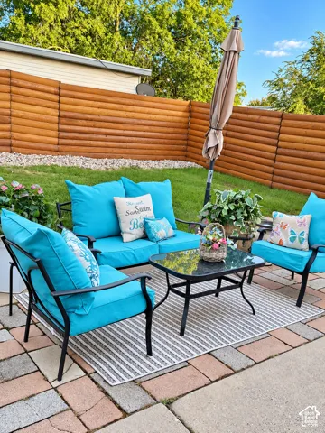 Outdoor entertaining space featuring a brick and stone patio, black metal frame lounge seating with cushions, glass-top cocktail table, mature landscaping with privacy fencing, and a cantilever umbrella for shade