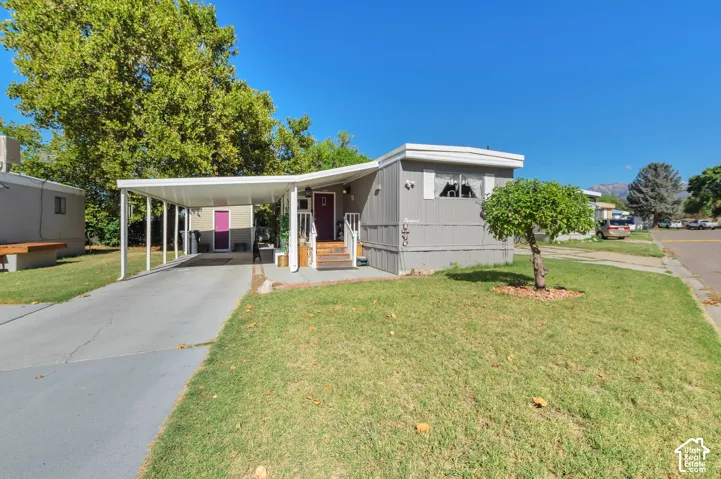Gray-sided manufactured home featuring a covered carport, front porch entry, and a manicured lawn