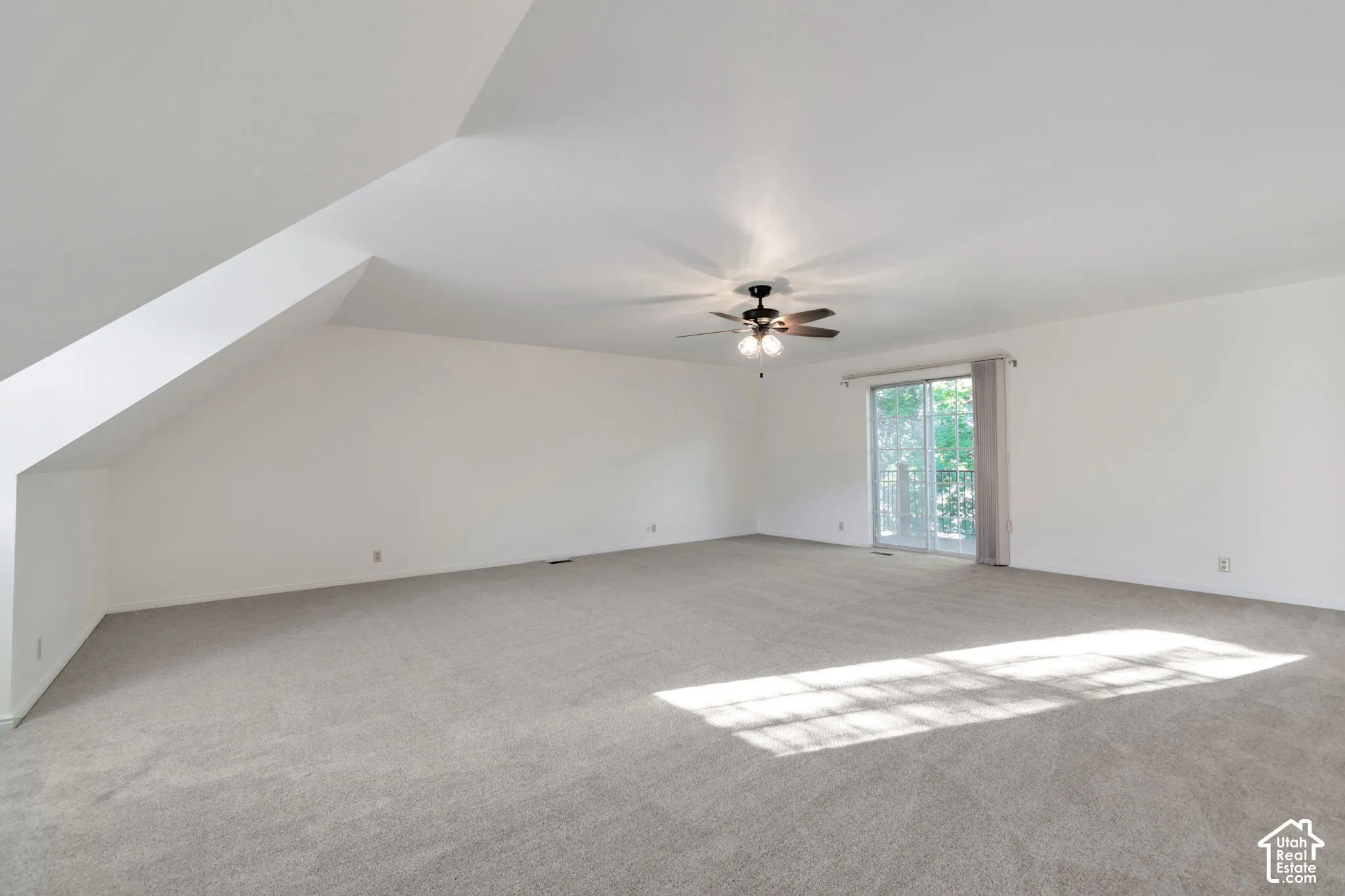 Spacious room featuring sloped ceilings, light-toned carpeting, a ceiling fan, and sliding glass doors
