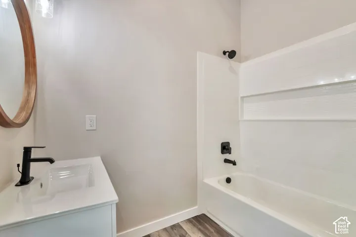 Wood-finish flooring, white vanity with integrated sink, black modern faucet, round wood-framed mirror, and white tiled tub surround with recessed shelf