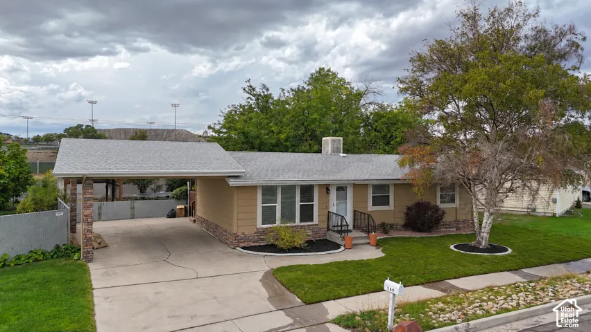 Carport with stone-clad support columns, concrete driveway, front entrance with handrail, landscaped front yard with mature trees, and a mail post