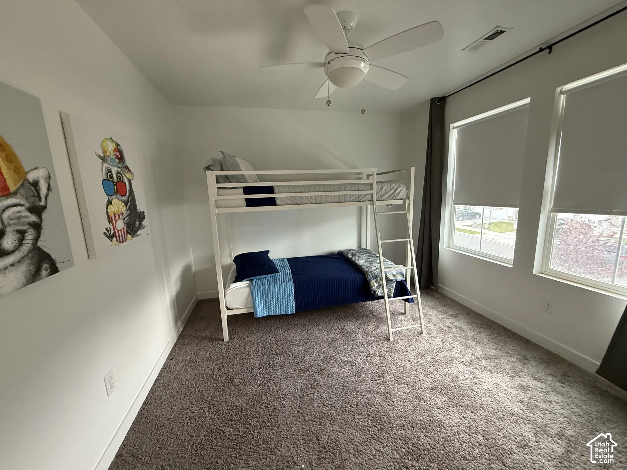 Plush carpet flooring, multiple windows with roller shades, white walls, and a ceiling fan