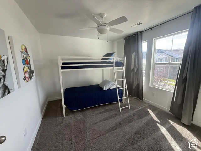 White walls, ceiling fan, large window, and dark carpet flooring