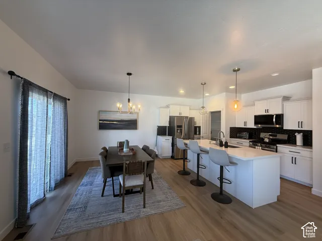Open-concept living space with wood-finish flooring, a contemporary kitchen featuring a large island with seating, pendant lighting, white cabinetry, and stainless steel appliances
