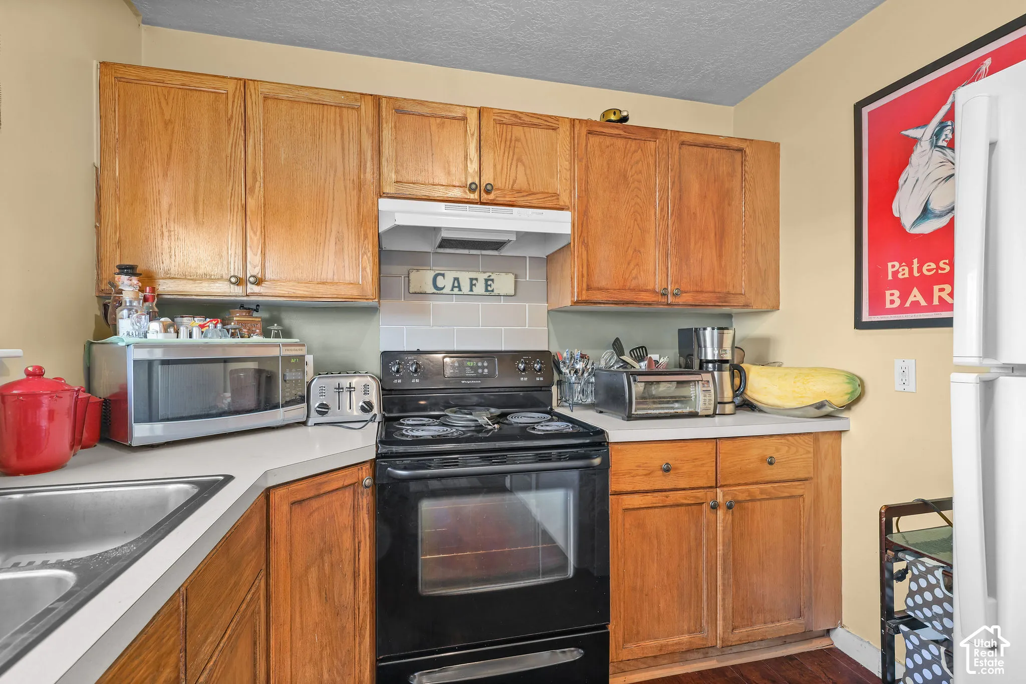 Unit B, Wood-finish cabinetry, light-colored countertops, stainless steel sink, black range, and a tiled backsplash