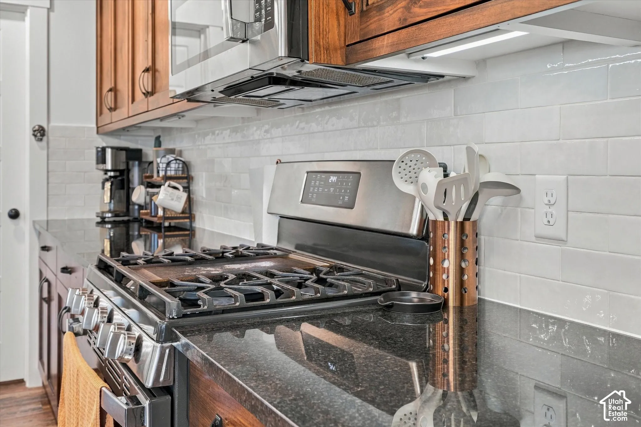 Stainless steel gas range with a griddle, black countertop, white subway tile backsplash, under-cabinet lighting, and wood cabinetry