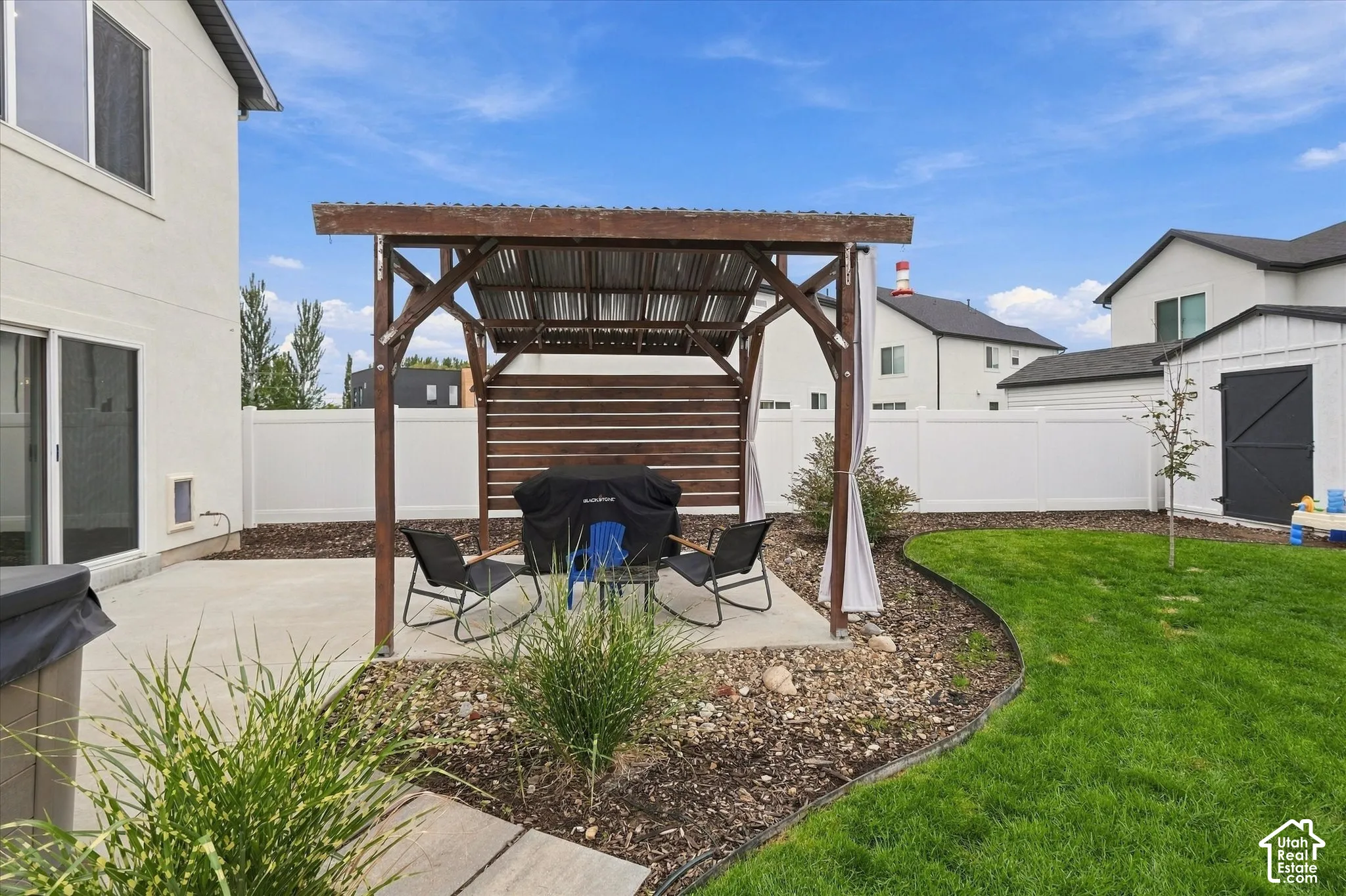 Expansive yard featuring a concrete patio, a wood-framed pergola with a corrugated roof, a manicured lawn, and a white privacy fence