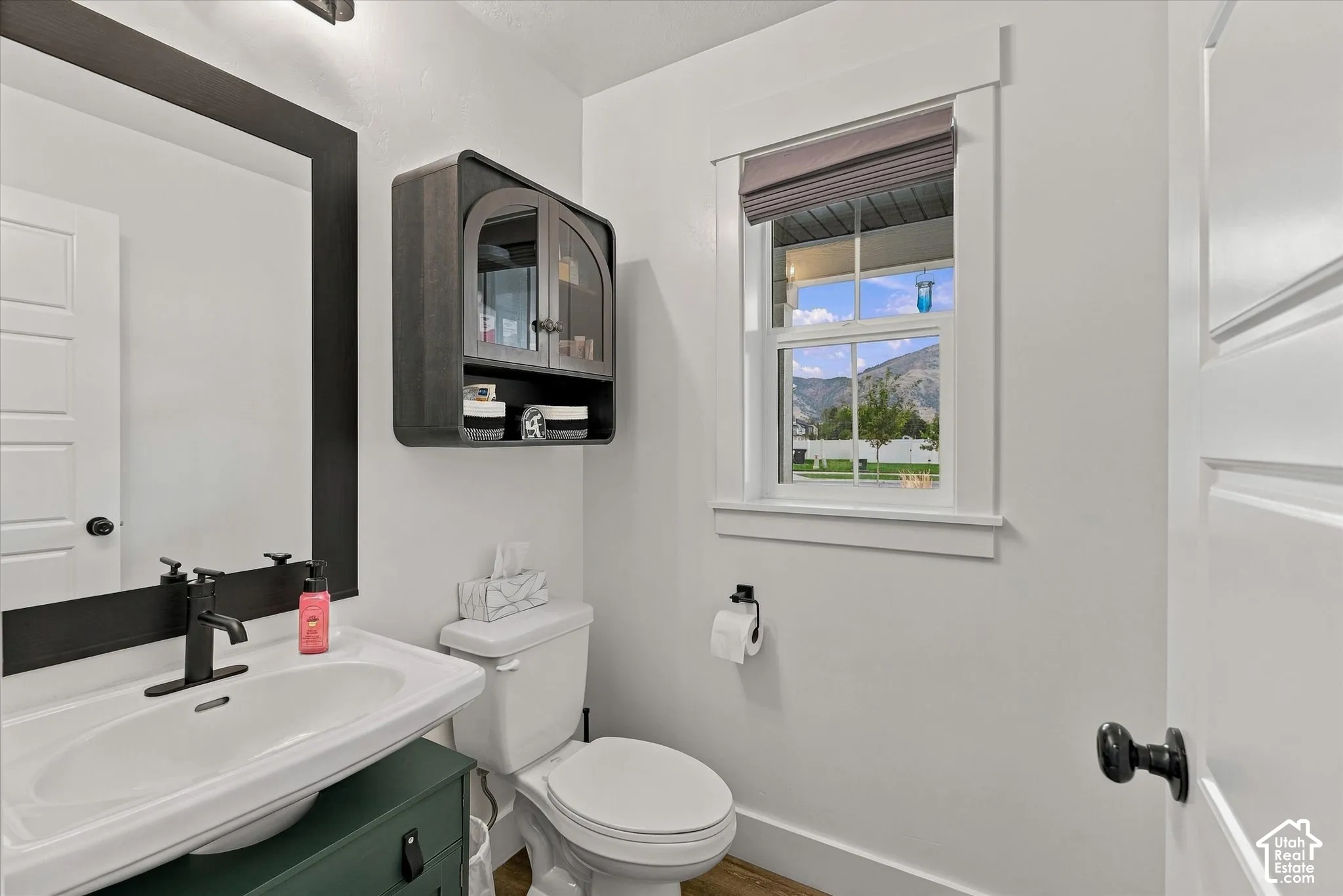 White paneled door with black hardware, black-framed mirror, wall-mounted storage cabinet, window with white trim, and a white toilet