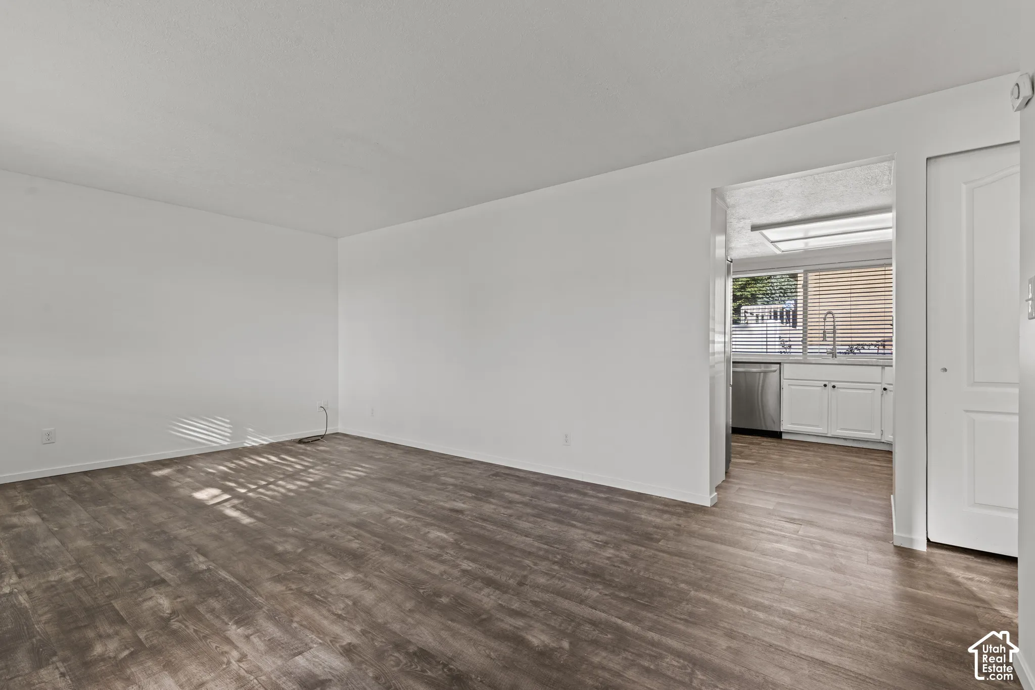 Wood-finish flooring, white walls, and a view into the kitchen featuring white cabinetry, a stainless steel dishwasher, and a window with horizontal blinds