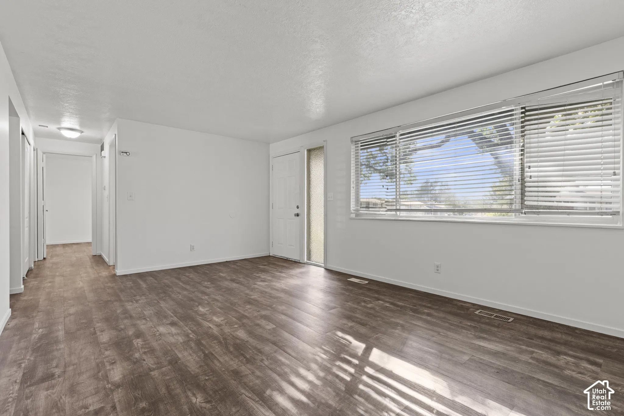 Spacious living area featuring wood-finish flooring, a large window with horizontal blinds, and a white entry door