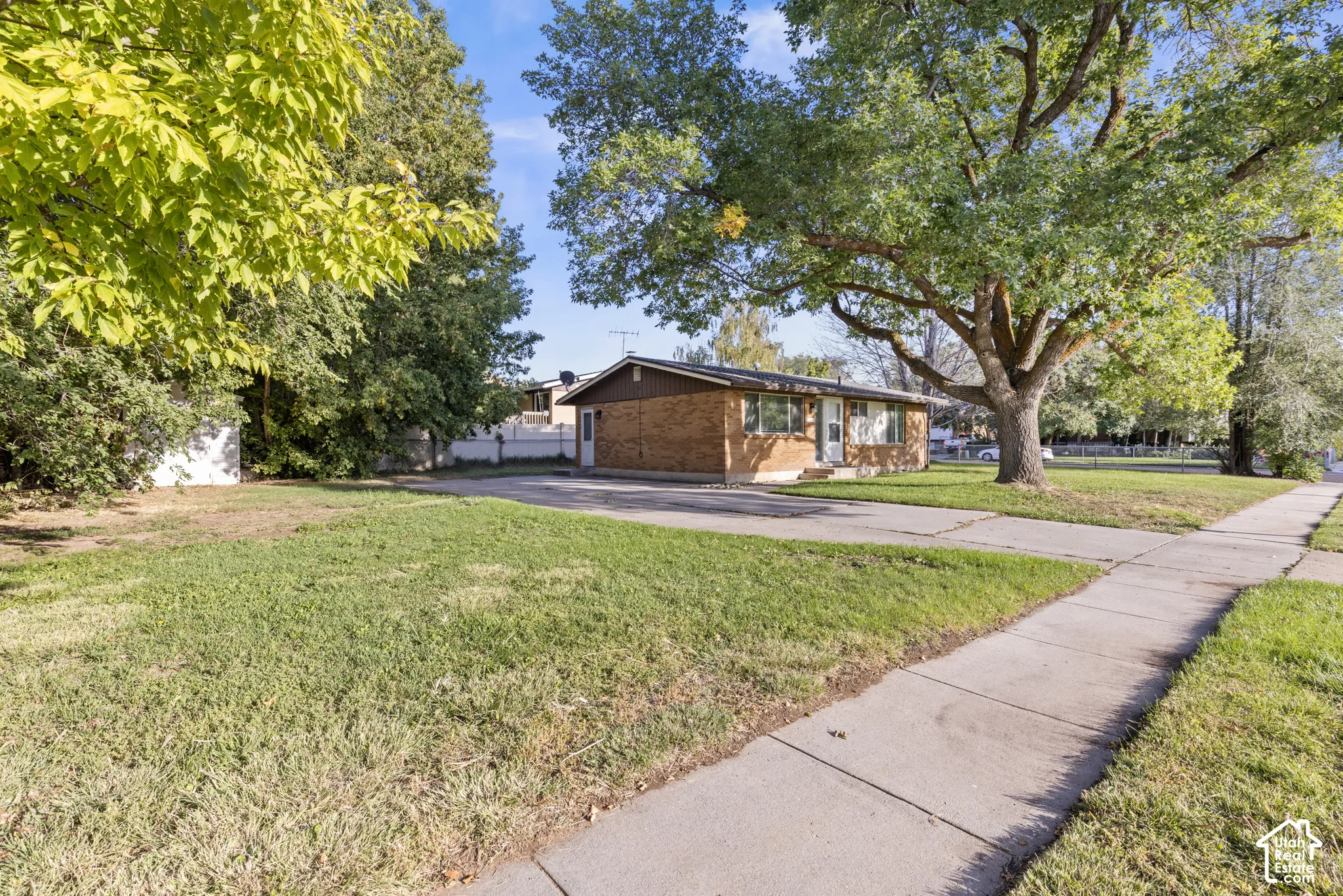 Brown brick exterior with a dark roof, front lawn, mature trees, and a concrete sidewalk
