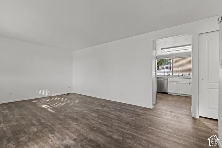 Wood-finish flooring, white walls, and a view into the kitchen featuring white cabinetry, a stainless steel dishwasher, and a window with horizontal blinds