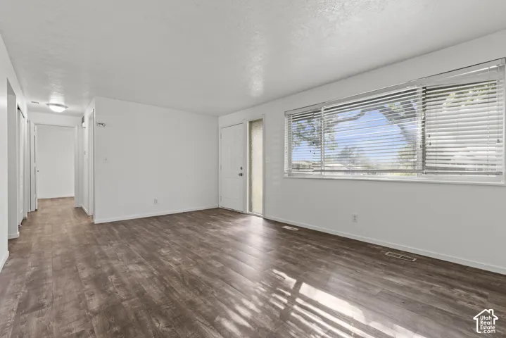 Spacious living area featuring wood-finish flooring, a large window with horizontal blinds, and a white entry door