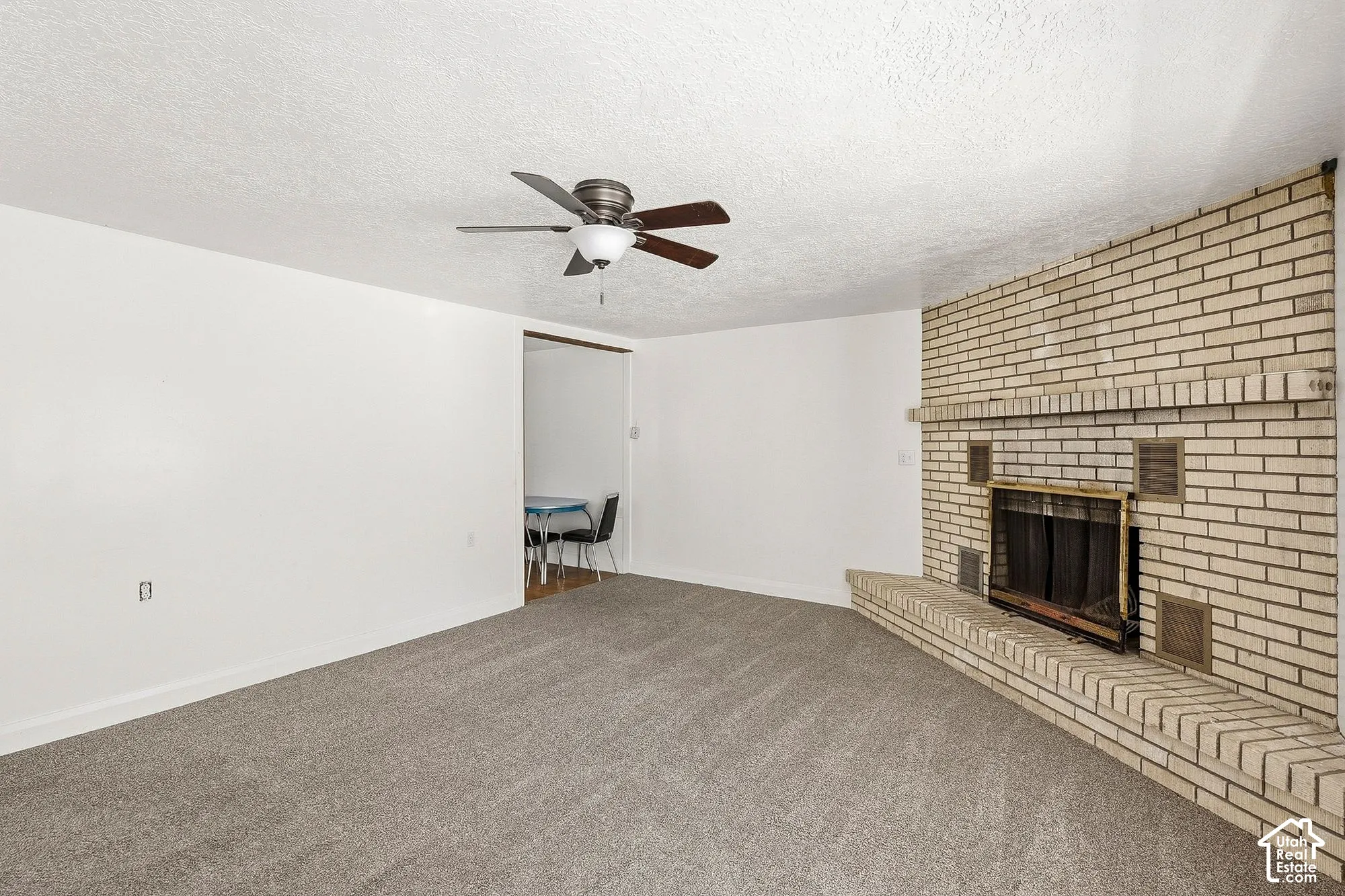 Light-colored brick fireplace with a hearth, ceiling fan, and grey carpet flooring