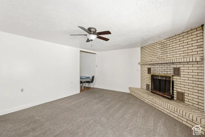 Light-colored brick fireplace with a hearth, ceiling fan, and grey carpet flooring