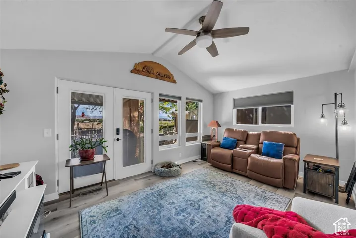 Vaulted ceiling, wood-finish flooring, ceiling fan, double glass-paneled doors, and multiple windows with roller shades
