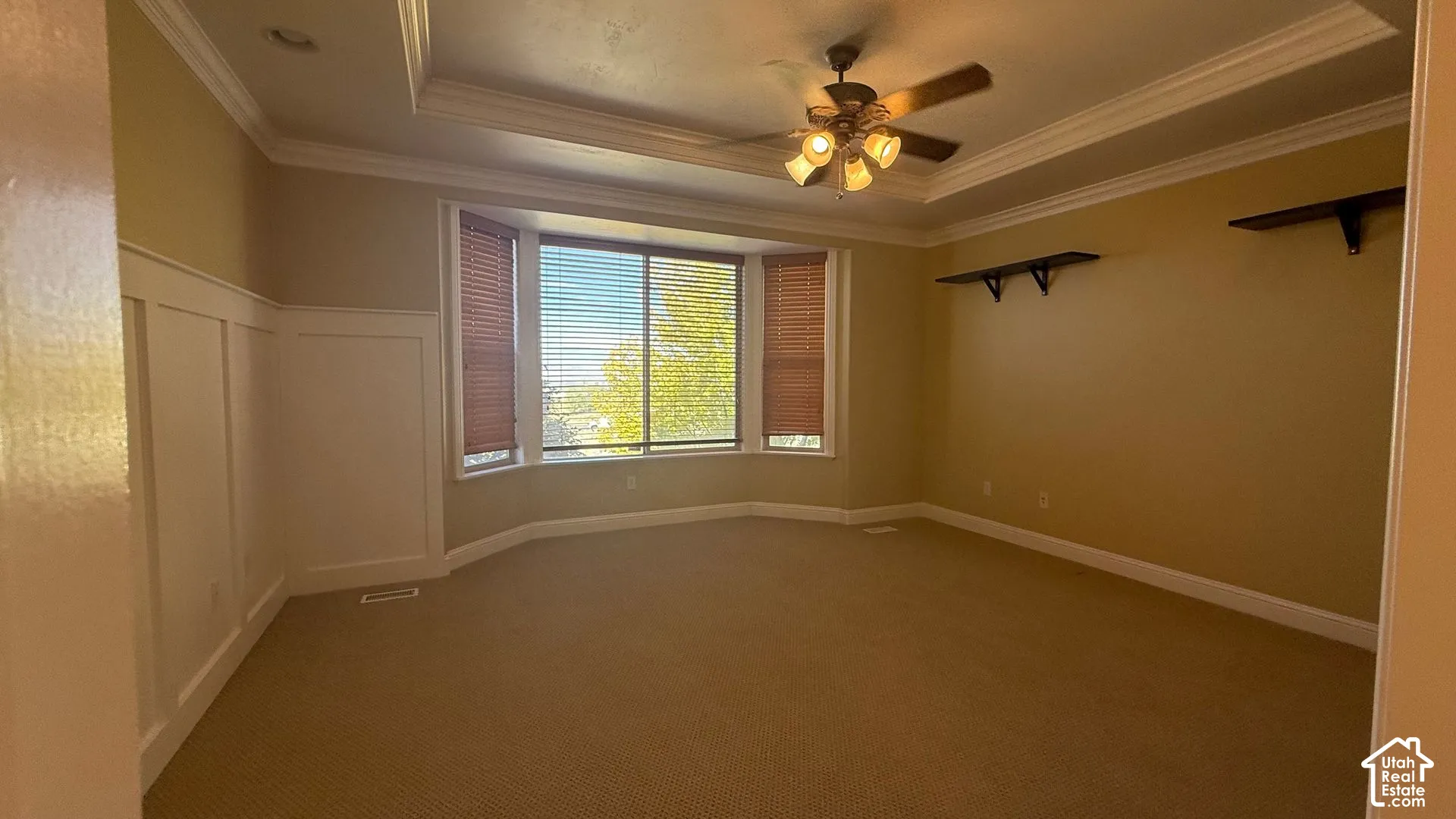 Recessed tray ceiling with crown molding, ceiling fan, bay window with blinds, wainscoting paneling, and wall-mounted shelving