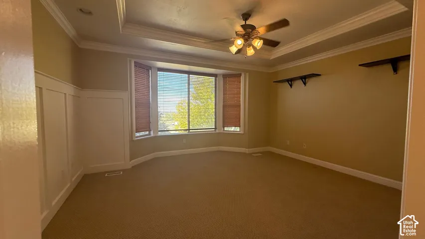 Recessed tray ceiling with crown molding, ceiling fan, bay window with blinds, wainscoting paneling, and wall-mounted shelving