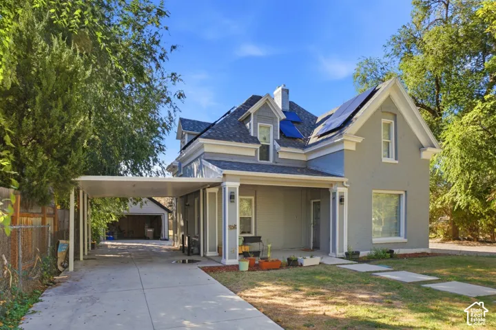 Gray exterior with white trim, covered front porch, integrated solar panels, paved driveway, and detached covered carport. Front of main house.