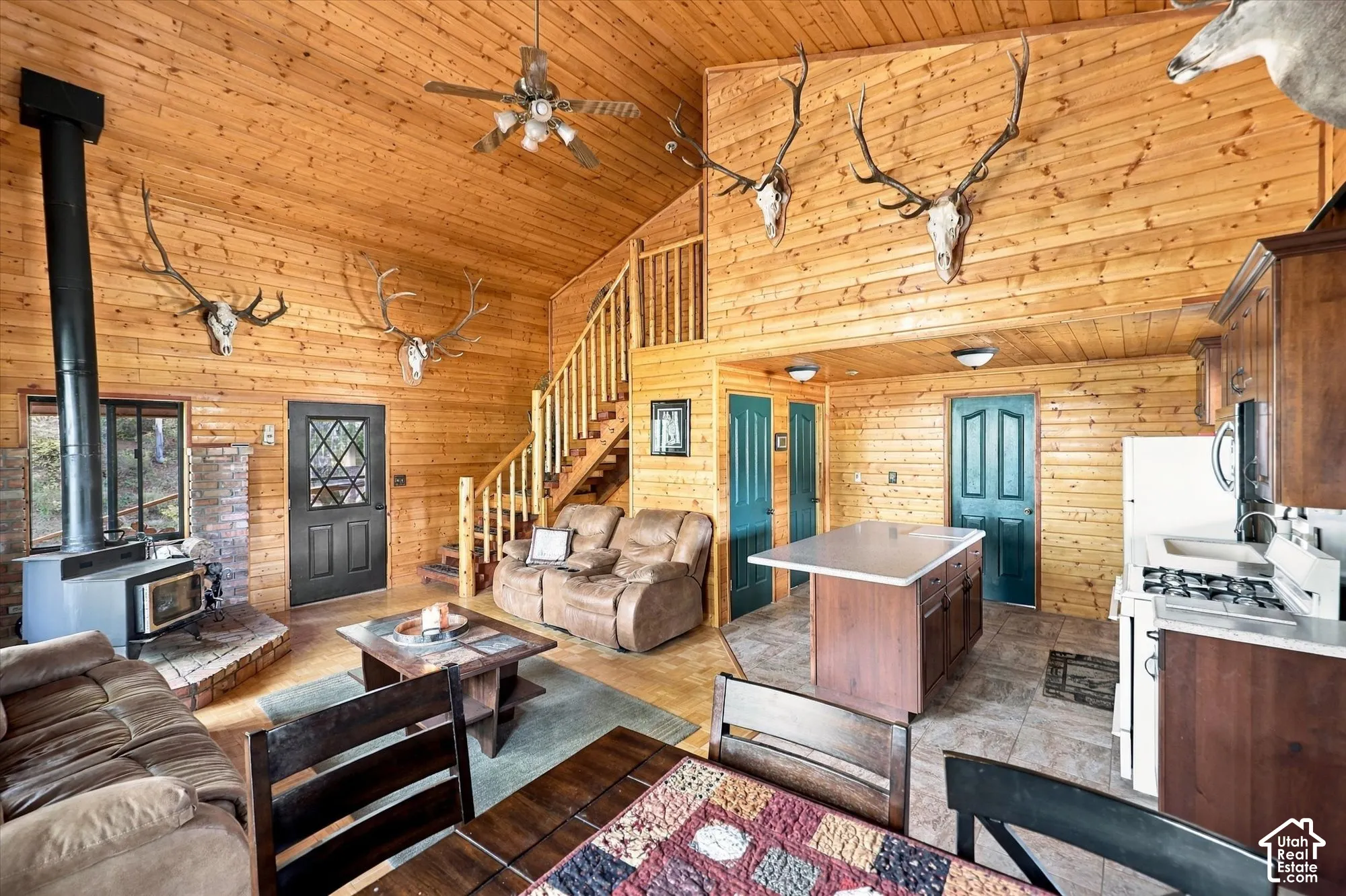 Vaulted wood-paneled ceilings with exposed beams, central wood-burning stove with brick hearth, open-concept kitchen with island, wood cabinetry, and gas range