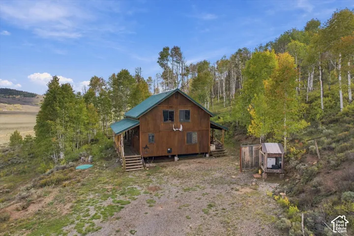 Wood-sided exterior with a green metal roof, elevated deck, and a natural, sloped landscape featuring mature trees