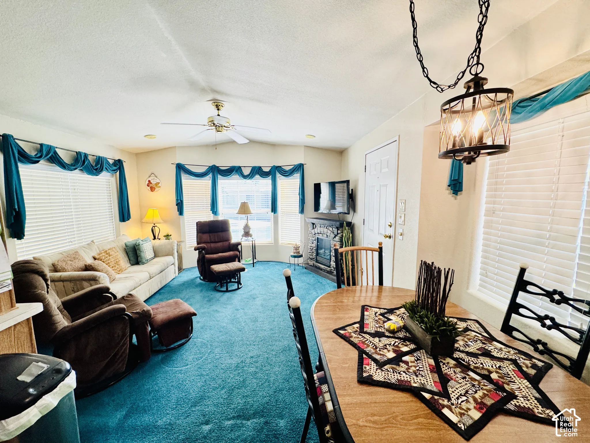 Open-concept living space with blue carpeting, a stone-front fireplace, a ceiling fan, and a dining area with a wrought iron chandelier