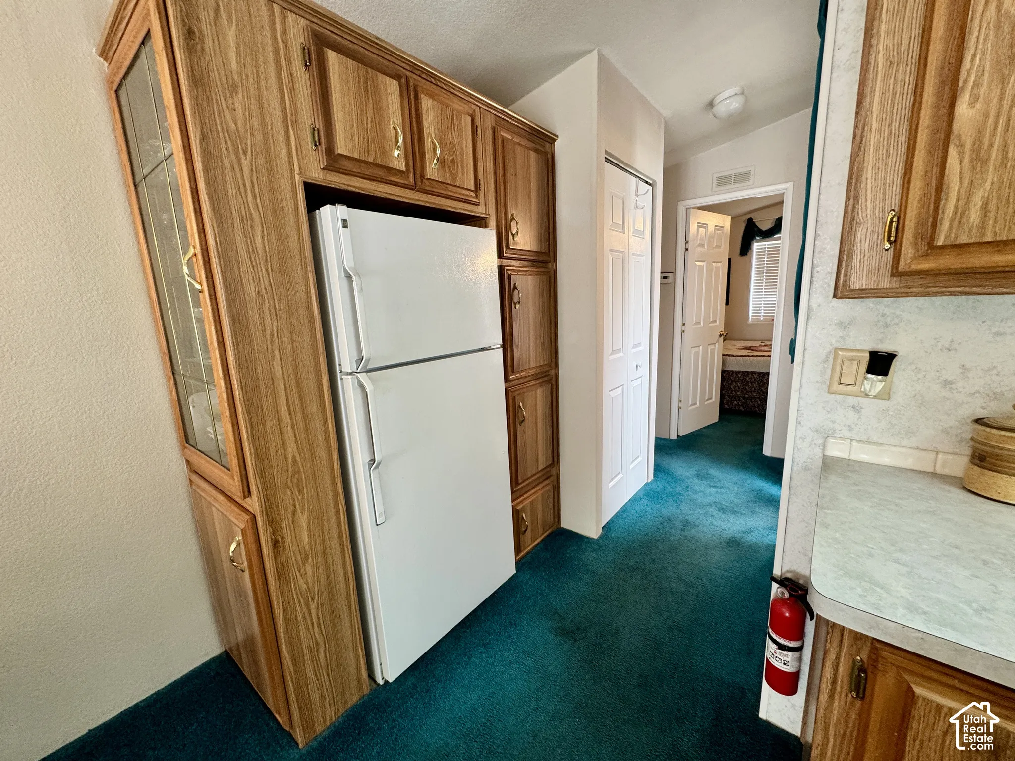 Wood-finish cabinetry with glass-front display, white freestanding refrigerator, teal carpet flooring, and white bifold closet doors