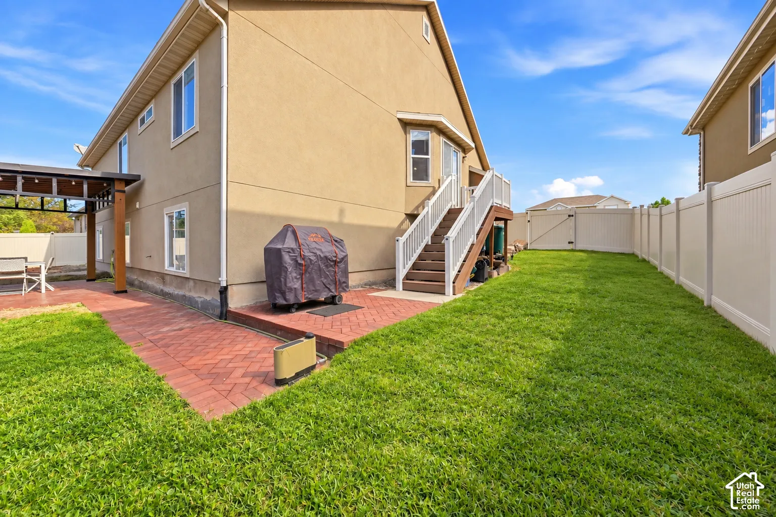 Expansive backyard featuring a manicured lawn bordered by vinyl fencing, brick patio, and detached two-story residence with staircase entry, covered deck, and tan exterior finish