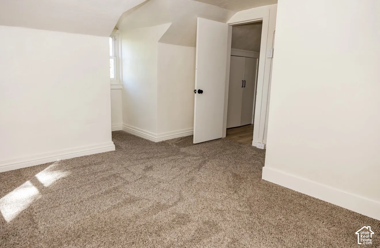 Light-toned carpet flooring, white painted walls, white trim, attic ceiling, and a white interior door with black hardware