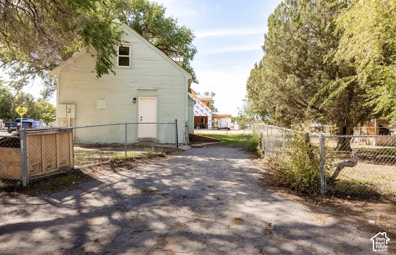 Light green siding exterior with a white entry door, chain-link fencing, mature trees, and a gravel drive