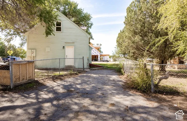 Light green siding exterior with a white entry door, chain-link fencing, mature trees, and a gravel drive