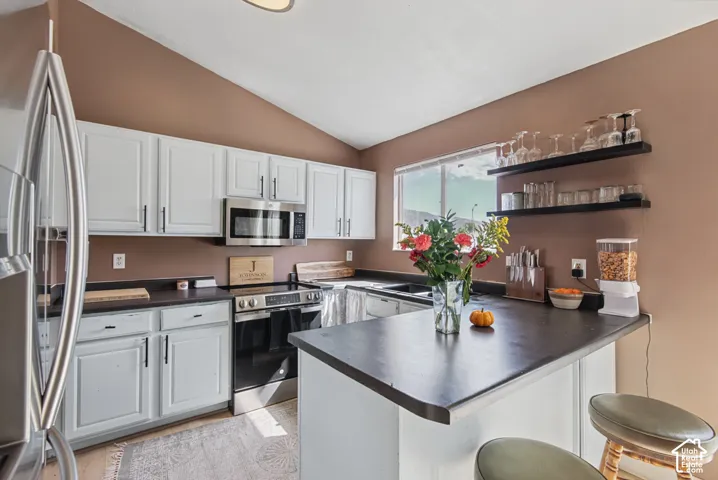 Vaulted ceiling kitchen featuring white cabinetry, stainless steel appliances, dark countertops, and a built-in breakfast bar