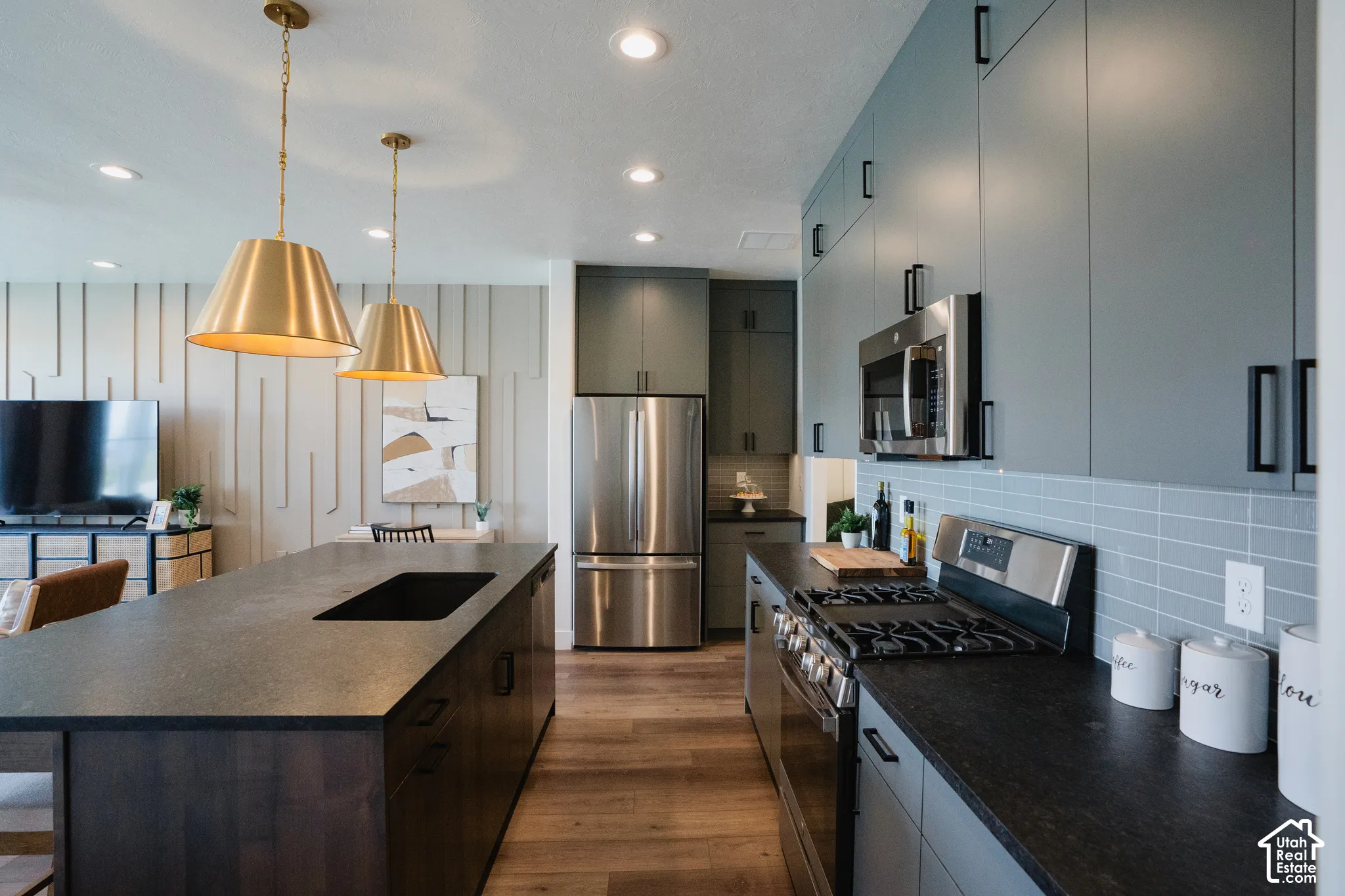 Kitchen featuring a dark-toned island with an integrated cooktop, brass pendant lighting, sleek flat-panel cabinetry, a light blue subway tile backsplash, and stainless steel appliances