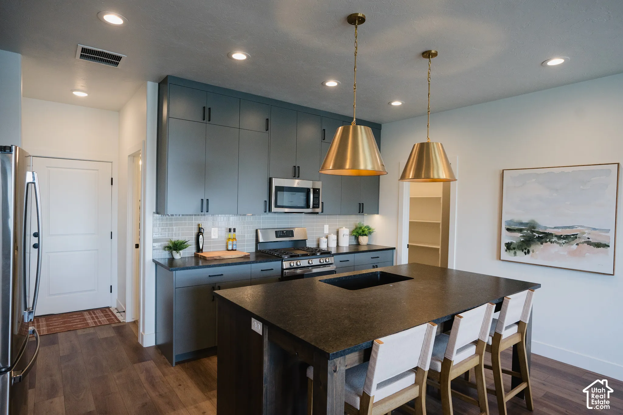 Kitchen featuring dark grey cabinetry, stainless steel appliances, a tile backsplash, wood-finish flooring, and a large island with integrated sink