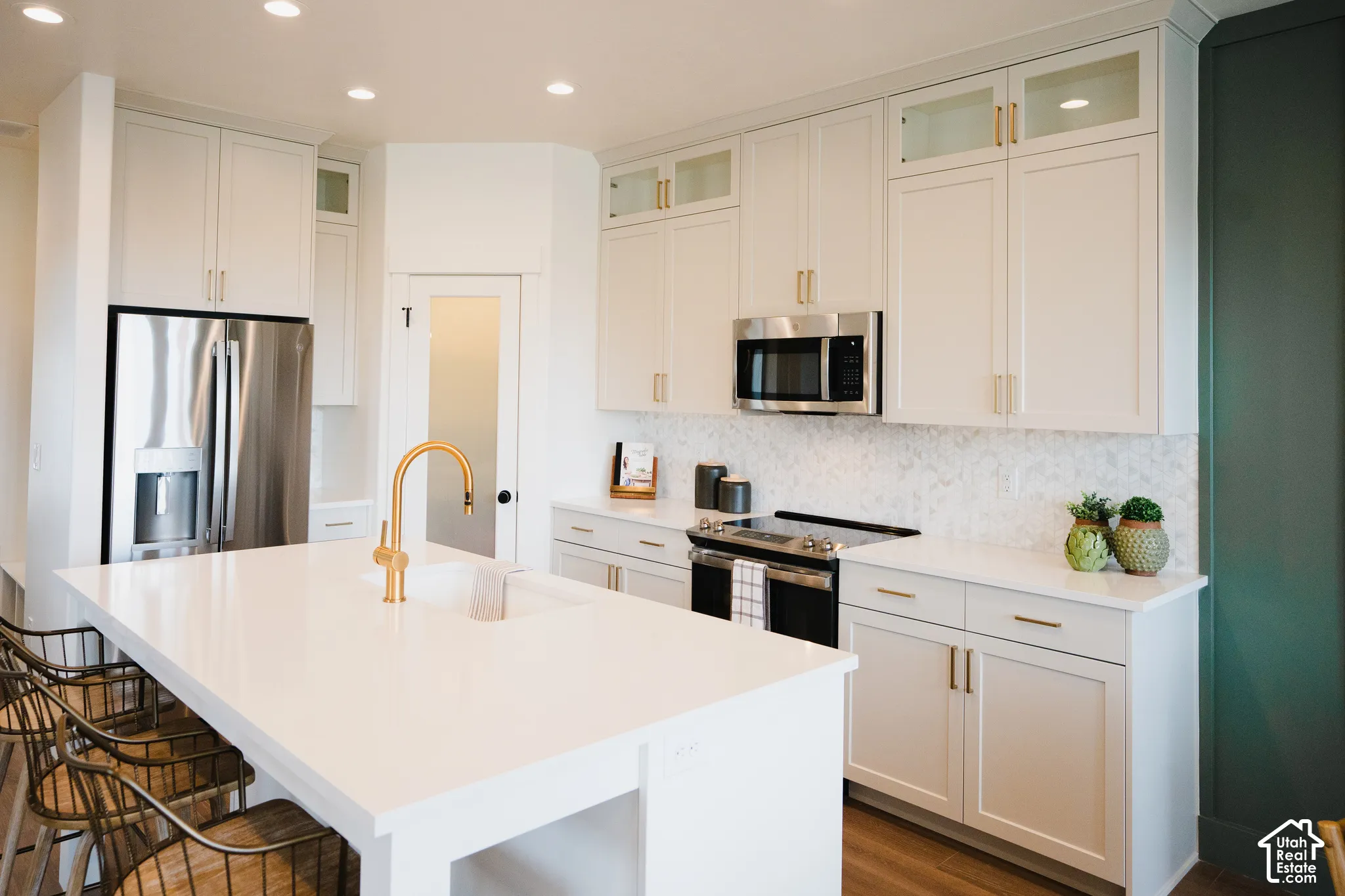 Kitchen featuring a white countertop island with integrated sink, stainless steel appliances, white cabinetry with gold-toned hardware, and a light-toned backsplash